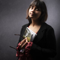 A portrait of a woman in a sweater holding a book and a pair of glasses
