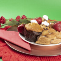 A plate with biscuits, muffins and strawberries on top on a bronze charger on a doily in front of a green background with the largest muffin in focus.