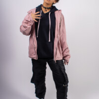 A model posing with a right hand up and left foot forward. Wearing a pink jacket with all black clothing underneath