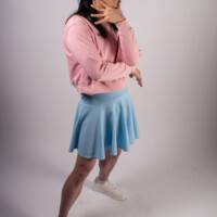 A model in a pink sweater and light blue skirt, posing to the side with a hand by their chin.
