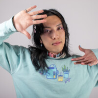 A model in a light blue sweater with light blue and pink accessories posing with their right hand by their face and left hand by their shoulder.