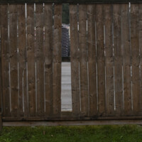 A photo of a backyard fence with a board missing and a parking lot behind on an overcast day.